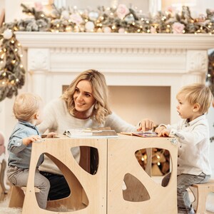 May include: A woman and two children sit at a wooden table with cut-out shapes. The table is made of light wood and has a natural finish. The children are sitting on the built-in stools. The woman is smiling and looking at the children. The children are looking at each other. The table is set with a book on it.