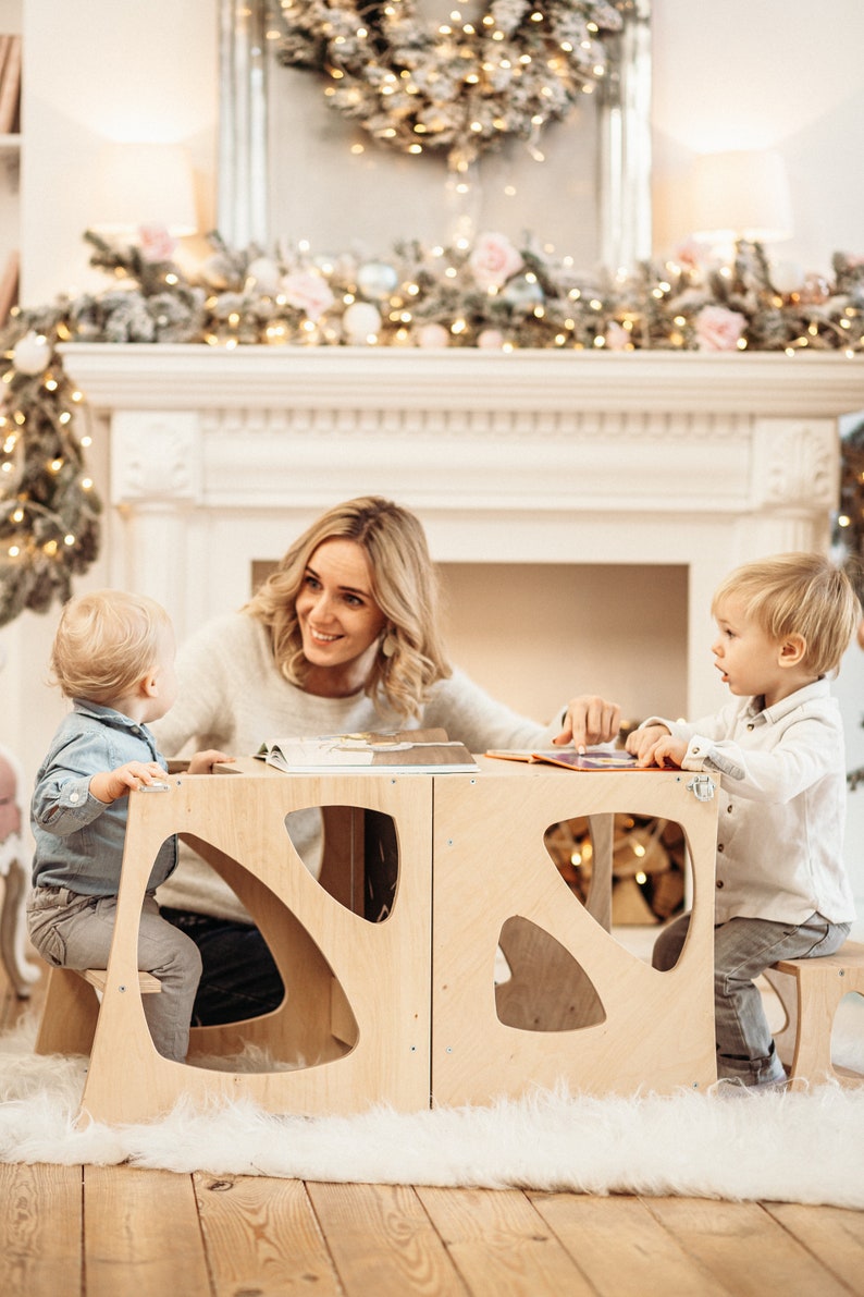 May include: A woman and two children sit at a wooden table with cut-out shapes. The table is made of light wood and has a natural finish. The children are sitting on the built-in stools. The woman is smiling and looking at the children. The children are looking at each other. The table is set with a book on it.