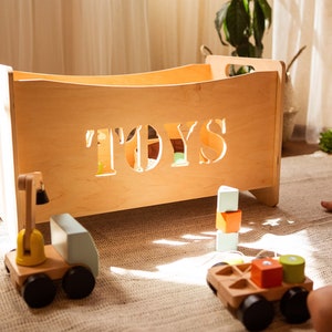 May include: A wooden toy box with the word "TOYS" cut out on the front. The box is filled with wooden toys, including a crane truck, a flatbed truck, and colourful blocks. The toys are on a rug.