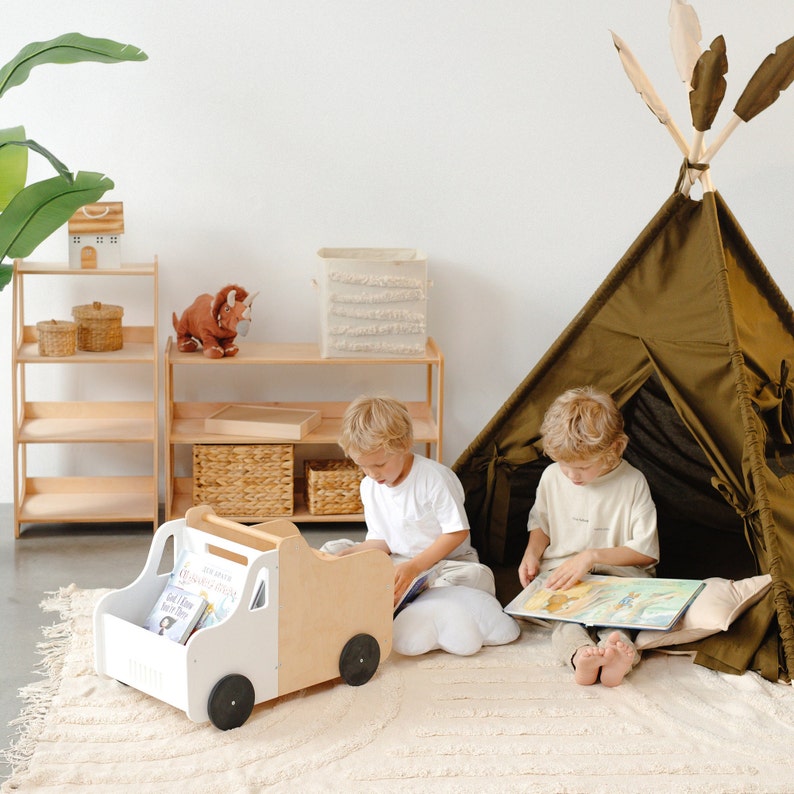Puede incluir: Dos ni&ntilde;os est&aacute;n sentados en una alfombra beige leyendo libros en una sala de juegos. Un cami&oacute;n de juguete de madera con libros dentro est&aacute; en la alfombra. Una tienda de campa&ntilde;a tipi est&aacute; en el fondo.
