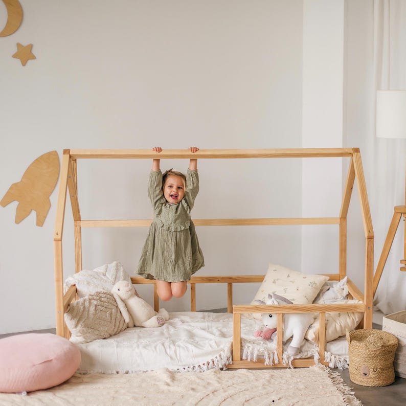 May include: A wooden house-shaped bed frame with a white mattress and a young girl hanging from the top rail. The bed is decorated with pillows and stuffed animals.