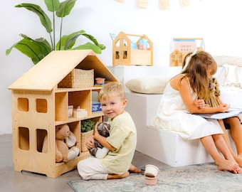 Puede incluir: Una casa de muñecas de madera con múltiples estantes, llena de juguetes y objetos decorativos. Un niño pequeño sostiene un peluche frente a la casa de muñecas. Otros dos niños están sentados cerca, leyendo un libro. La casa de muñecas es de madera clara.