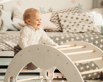 May include: A white wooden climbing arch with a toddler climbing on it. The arch has a rounded top and several horizontal rungs. The toddler is wearing a white shirt and grey pants. The arch is on a white rug in a room with a white wall and a grey and white patterned bed.