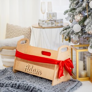 K&ouml;nnte beinhalten: Ein Babybett aus Holz mit einem roten Band und dem Namen "Adam" auf der Vorderseite eingraviert. Das Bett steht auf einer grauen Strickdecke vor einem Weihnachtsbaum.