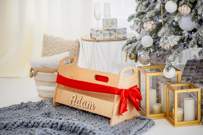 K&ouml;nnte beinhalten: Ein Babybett aus Holz mit einem roten Band und dem Namen "Adam" auf der Vorderseite eingraviert. Das Bett steht auf einer grauen Strickdecke vor einem Weihnachtsbaum.