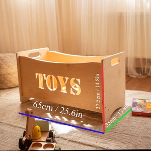 May include: A light-coloured wooden toy box with the word "TOYS" cut out on the front. The box is 65cm long, 37.5cm high, and 35cm wide. It has handles on the sides.