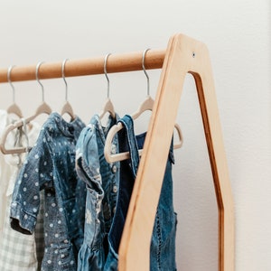 May include: A light-colored wooden clothing rack with an A-frame design, featuring a horizontal rod holding several small hangers with various garments, including a white checkered dress, a blue polka dot dress, and a blue denim overall dress.