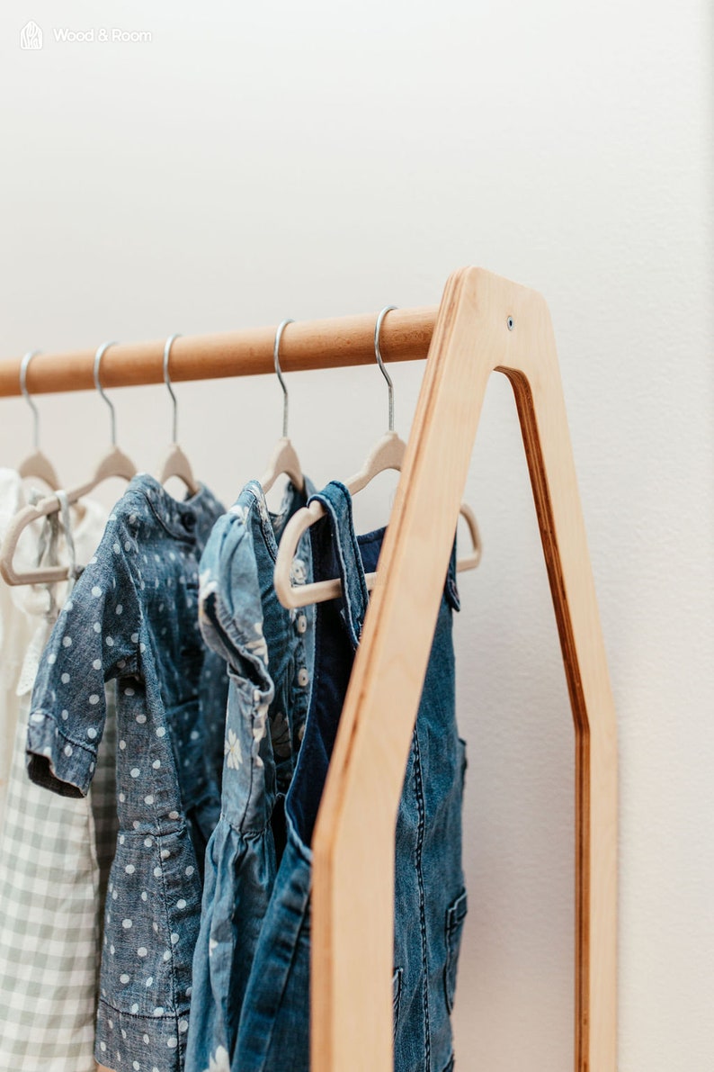 May include: A light-colored wooden clothing rack with an A-frame design, featuring a horizontal rod holding several small hangers with various garments, including a white checkered dress, a blue polka dot dress, and a blue denim overall dress.