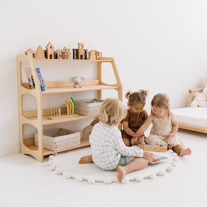 Könnte beinhalten: Drei Kinder spielen mit Holzspielzeug auf einem weißen, flauschigen Teppich vor einem Holzregal. Das Regal hat drei Regalbretter und ist mit Spielzeug gefüllt, darunter ein Holzelefant, ein Regenbogen-Abakus und ein Buch mit dem Titel "Little Blue Truck".
