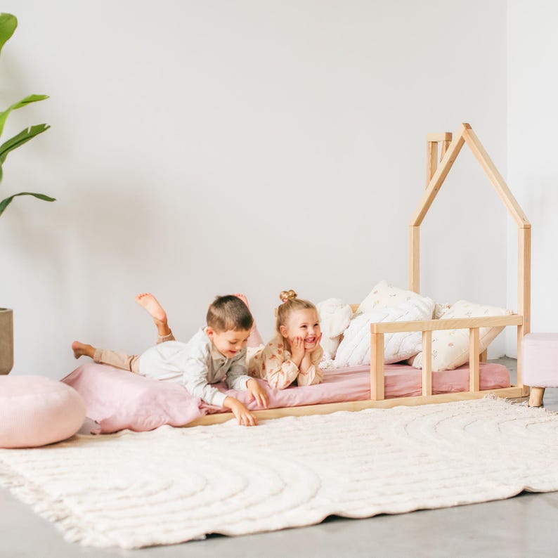 May include: Two children are lying on a pink bed with a wooden house-shaped frame. The bed is on a white fluffy rug. The children are smiling and looking at each other.