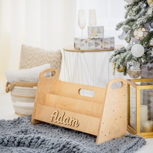 K&ouml;nnte beinhalten: Ein B&uuml;cherregal aus Holz mit dem Namen "Adam" auf der Vorderseite. Das Regal hat zwei Regalbretter und ist aus hellem Holz gefertigt. Es steht auf einer grauen Strickdecke vor einem Weihnachtsbaum.