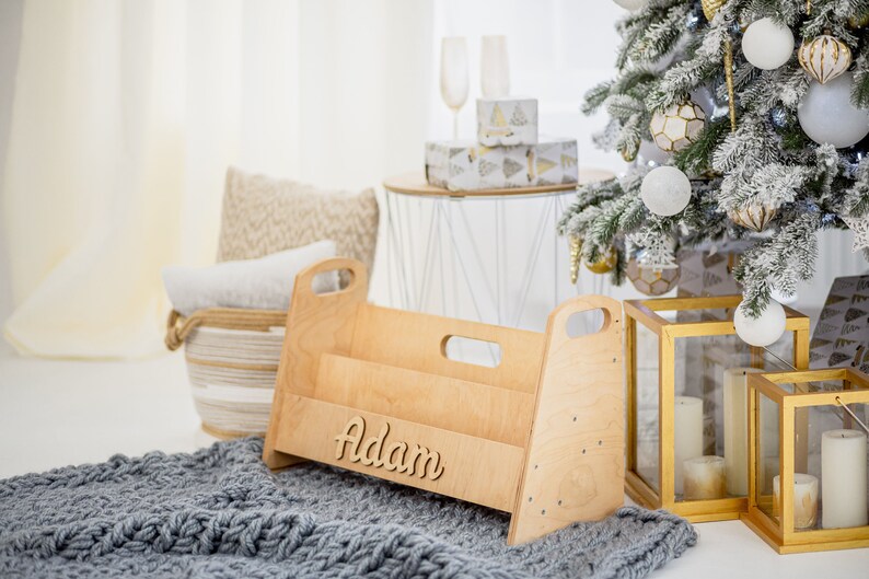 K&ouml;nnte beinhalten: Ein B&uuml;cherregal aus Holz mit dem Namen "Adam" auf der Vorderseite. Das Regal hat zwei Regalbretter und ist aus hellem Holz gefertigt. Es steht auf einer grauen Strickdecke vor einem Weihnachtsbaum.