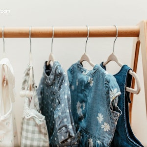 May include: A light-colored wooden clothing rack with a triangular frame holds six garments on metal hangers. The items include white ruffled tops, a light green checkered piece, a blue polka-dot shirt, and blue dresses with white floral patterns.