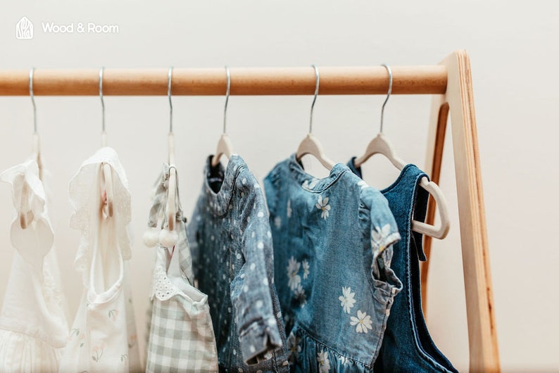 May include: A light-colored wooden clothing rack with a triangular frame holds six garments on metal hangers. The items include white ruffled tops, a light green checkered piece, a blue polka-dot shirt, and blue dresses with white floral patterns.
