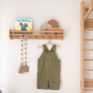 Pu&ograve; includere: Uno scaffale in legno con un libro intitolato "Little Blue Truck" e un set di archi in legno arcobaleno. Un completo verde da lavoro &egrave; appeso a un appendiabiti in legno sotto lo scaffale. Una ghirlanda di perline bianche con nappe &egrave; appesa allo scaffale.