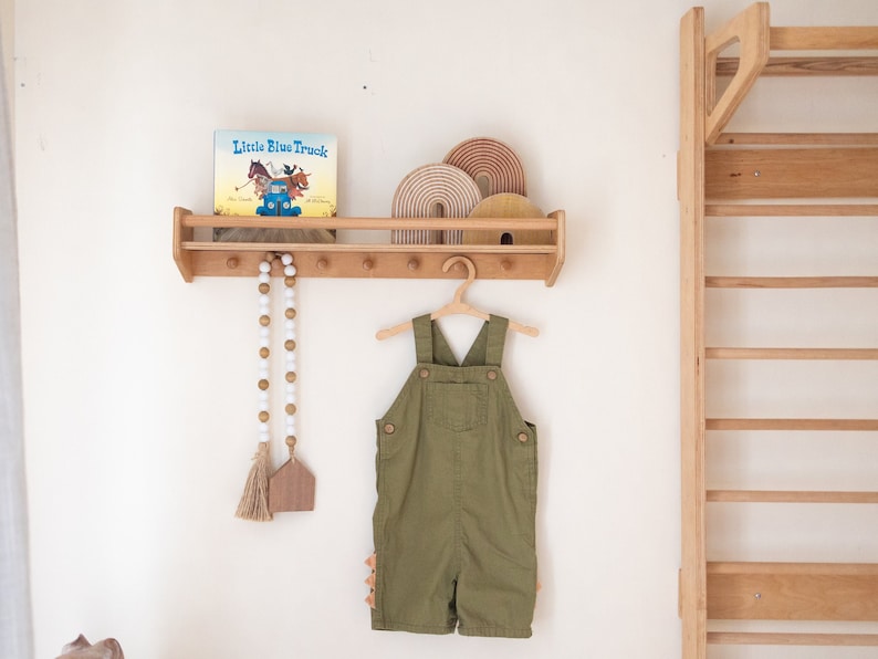 Pu&ograve; includere: Uno scaffale in legno con un libro intitolato "Little Blue Truck" e un set di archi in legno arcobaleno. Un completo verde da lavoro &egrave; appeso a un appendiabiti in legno sotto lo scaffale. Una ghirlanda di perline bianche con nappe &egrave; appesa allo scaffale.
