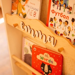 May include: Wooden bookshelf with the name "emma" in white script. The shelf holds several children's books with colorful covers, including one with a cartoon lion and another with the word "Words".