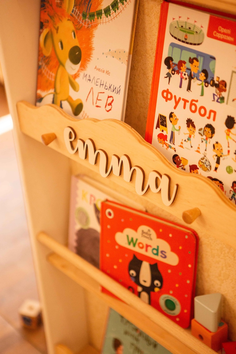 May include: Wooden bookshelf with the name "emma" in white script. The shelf holds several children's books with colorful covers, including one with a cartoon lion and another with the word "Words".