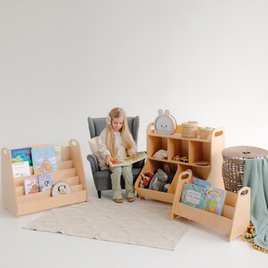 K&ouml;nnte beinhalten: Ein Kind sitzt in einem grauen Sessel und liest ein Buch. Es gibt drei Holzbuchregale mit Regalen zum Verstauen von B&uuml;chern und Spielzeug. Die B&uuml;cherregale sind in hellem Holz gehalten. Die B&uuml;cherregale haben einen Griff an der Seite. Die B&uuml;cherregale stehen auf einem hellbeigen Teppich.