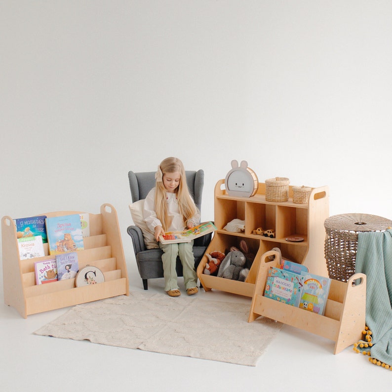 K&ouml;nnte beinhalten: Ein Kind sitzt in einem grauen Sessel und liest ein Buch. Es gibt drei Holzbuchregale mit Regalen zum Verstauen von B&uuml;chern und Spielzeug. Die B&uuml;cherregale sind in hellem Holz gehalten. Die B&uuml;cherregale haben einen Griff an der Seite. Die B&uuml;cherregale stehen auf einem hellbeigen Teppich.