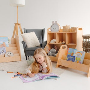 K&ouml;nnte beinhalten: Ein junges M&auml;dchen sitzt auf dem Boden mit einem Buch, das vor ihr offen liegt. Sie ist umgeben von Holzbuchregalen, die mit bunten Kinderb&uuml;chern gef&uuml;llt sind. Die B&uuml;cherregale sind in hellem Holz gehalten und haben ein schlichtes Design. Das M&auml;dchen tr&auml;gt ein wei&szlig;es Hemd und Jeans.