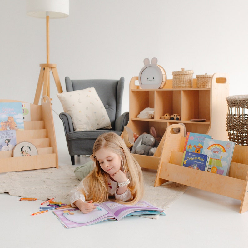 K&ouml;nnte beinhalten: Ein junges M&auml;dchen sitzt auf dem Boden mit einem Buch, das vor ihr offen liegt. Sie ist umgeben von Holzbuchregalen, die mit bunten Kinderb&uuml;chern gef&uuml;llt sind. Die B&uuml;cherregale sind in hellem Holz gehalten und haben ein schlichtes Design. Das M&auml;dchen tr&auml;gt ein wei&szlig;es Hemd und Jeans.