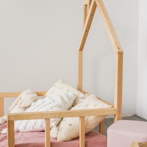 May include: A wooden toddler bed with a house-shaped frame. The bed has a pink sheet and several white and cream pillows. A pink footstool and a woven basket are in the foreground.