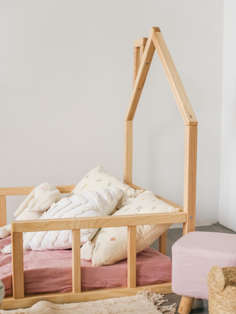 May include: A wooden toddler bed with a house-shaped frame. The bed has a pink sheet and several white and cream pillows. A pink footstool and a woven basket are in the foreground.