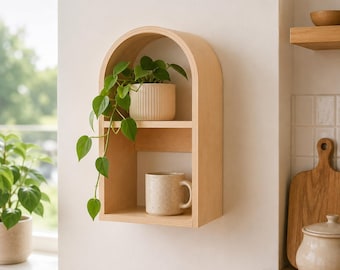 Op de afbeelding: Een lichtgekleurd houten wandrek met een gewelfde bovenkant, met twee planken. Een potplant met groene bladeren staat op de bovenste plank en een gespikkelde mok staat op de onderste plank. Het rek is aan een witte muur bevestigd.