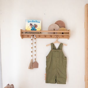 May include: A wooden shelf with a book titled "Little Blue Truck" and a wooden toy rainbow on top. There are wooden hooks on the shelf with a white beaded garland hanging from one of them. A green overall is hanging from a wooden hanger on the hooks.