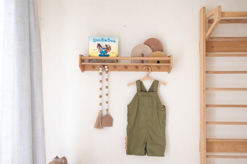 May include: A wooden shelf with a book titled "Little Blue Truck" and a wooden toy rainbow on top. There are wooden hooks on the shelf with a white beaded garland hanging from one of them. A green overall is hanging from a wooden hanger on the hooks.