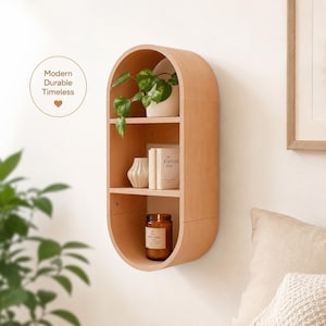 May include: A light-colored, oval-shaped wooden wall shelf with three shelves. The shelf holds a potted plant, books, a small vase, and a candle. The text "Modern Durable Timeless" is in a circle in the upper left corner.