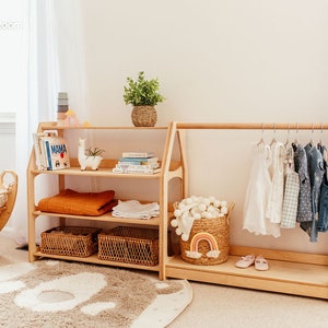 May include: Wooden children's room furniture set. Includes a three-shelf bookcase with books, blankets, and baskets, and a clothing rack with hanging clothes. A woven basket with a rainbow decor and a rug are also present.