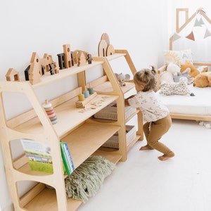 May include: A light wood bookshelf with five shelves, featuring a variety of wooden toys, including blocks, a stacking ring tower, and a wooden elephant. A young child is reaching for a toy on the top shelf.