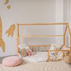 May include: A wooden toddler bed frame with a white sheet and pillows. The bed is decorated with stuffed animals and a pink throw pillow. The bed is in a room with white walls and a wooden floor.