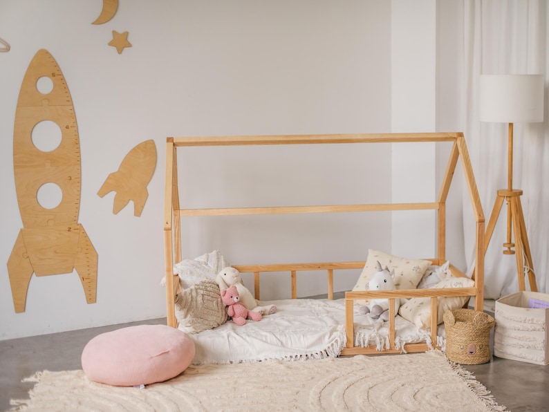 May include: A wooden toddler bed frame with a white sheet and pillows. The bed is decorated with stuffed animals and a pink throw pillow. The bed is in a room with white walls and a wooden floor.