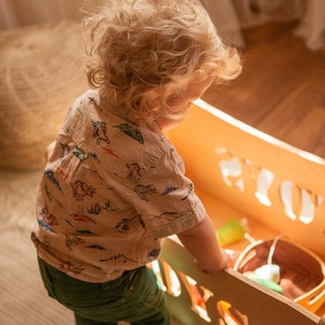 May include: A child in a dinosaur-print shirt and green trousers stands by a wooden toy box. The box has cut-out letters spelling a name. A woven pouffe is in the background. The scene is bathed in warm light.