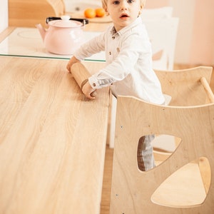 May include: A young child sits in a wooden learning tower, reaching for a rolling pin on a light wood countertop. The tower has a unique design with cutouts in the sides.