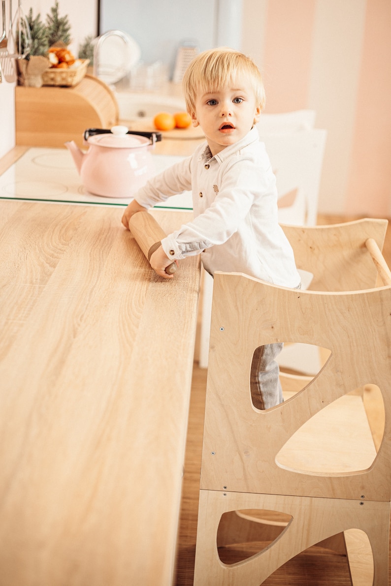 May include: A young child sits in a wooden learning tower, reaching for a rolling pin on a light wood countertop. The tower has a unique design with cutouts in the sides.