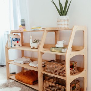 May include: A light wood, multi-shelf storage unit with a modern design. The shelves hold books, blankets, and decorative items, including a llama planter and a potted plant. Woven baskets are placed on the lower shelves. The unit is in a room with a rug.