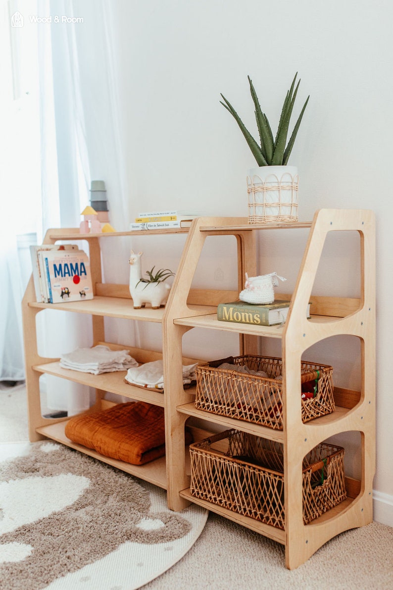 May include: A light wood, multi-shelf storage unit with a modern design. The shelves hold books, blankets, and decorative items, including a llama planter and a potted plant. Woven baskets are placed on the lower shelves. The unit is in a room with a rug.