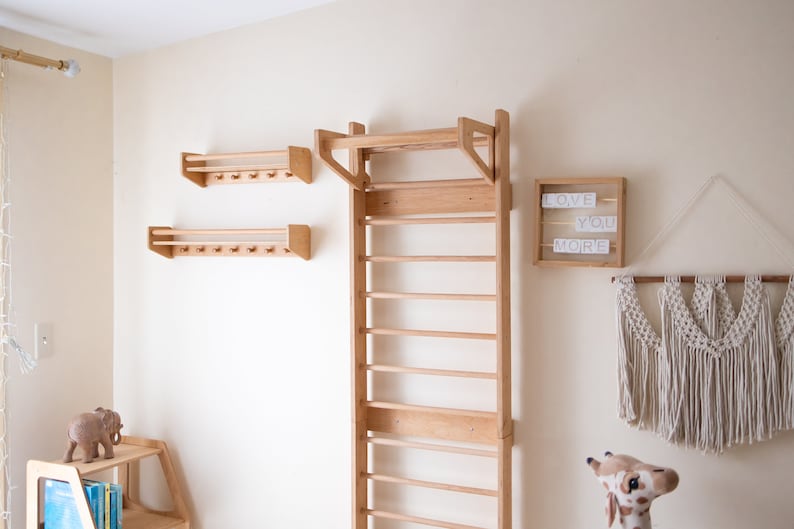 Pu&ograve; includere: Una scala da arrampicata in legno montata a parete con uno scaffale sopra. Ci sono due scaffali pi&ugrave; piccoli con ganci sotto la scala. Gli scaffali sono in legno chiaro e hanno una finitura naturale. C'&egrave; un cartello che dice "LOVE YOU MORE" sul muro a destra della scala.