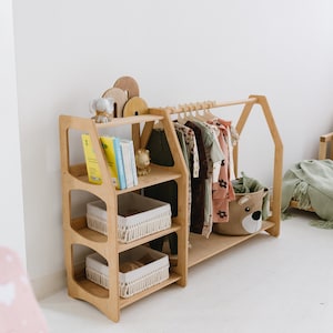 Op de afbeelding: Een vrijstaande kledingrek van hout met drie planken en een hangende stang. De planken hebben geweven manden voor opslag. Het rek is versierd met een klein olifantbeeldje en boeken. Er is een knuffeldier in een mand op de onderste plank.