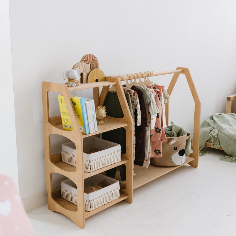 Op de afbeelding: Een vrijstaande kledingrek van hout met drie planken en een hangende stang. De planken hebben geweven manden voor opslag. Het rek is versierd met een klein olifantbeeldje en boeken. Er is een knuffeldier in een mand op de onderste plank.