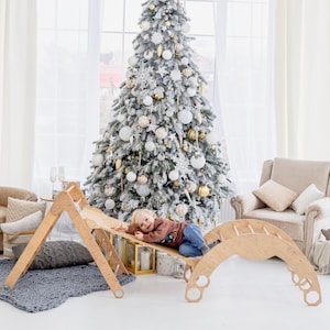 May include: A wooden climbing arch, ramp, and triangle set. The natural wood-toned play set is in a bright room with a decorated Christmas tree in the background. A child is lying on the ramp, smiling. The set is designed for indoor play.