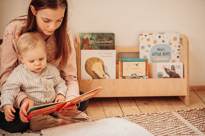 May include: A woman and a child sit on the floor reading a book together. The child is sitting on the woman's lap. The woman is holding the book open. The child is pointing at the book. There is a bookshelf behind them with several colorful children's books.
