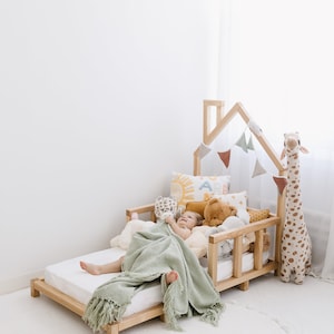 K&ouml;nnte beinhalten: Ein Kind schl&auml;ft in einem h&ouml;lzernen, hausf&ouml;rmigen Bett mit einer gr&uuml;nen Decke. Das Bett ist mit Stofftieren und einer Girlande aus Fahnen dekoriert. Das Bett steht auf einem wei&szlig;en Teppich mit einem wei&szlig;en Pompon-Saum.