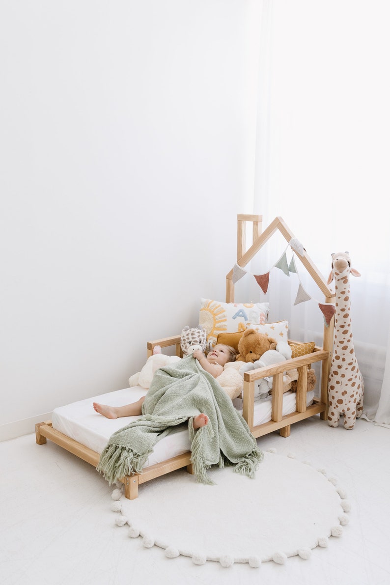 K&ouml;nnte beinhalten: Ein Kind schl&auml;ft in einem h&ouml;lzernen, hausf&ouml;rmigen Bett mit einer gr&uuml;nen Decke. Das Bett ist mit Stofftieren und einer Girlande aus Fahnen dekoriert. Das Bett steht auf einem wei&szlig;en Teppich mit einem wei&szlig;en Pompon-Saum.
