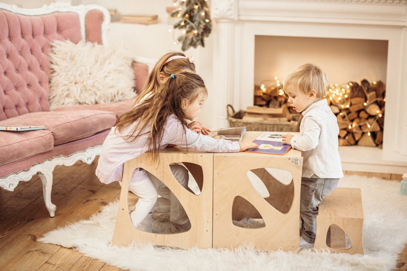 May include: Three children sitting at a wooden table with cutouts, reading a book together. The table is made of light wood and has a unique design. The children are sitting on stools and a step stool.