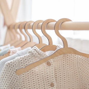 Op de afbeelding: Een close-up van een houten kledingrek met meerdere houten hangers met kleine gebreide kledingstukken. De hangers zijn van natuurlijk hout met een gladde afwerking.